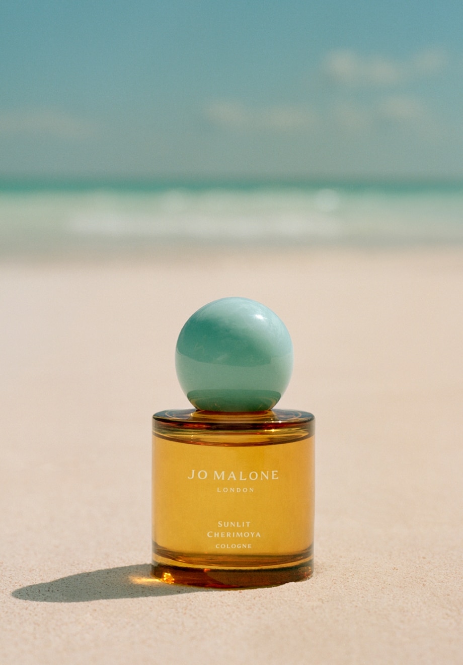 Jo Malone London Starlit Cherimoya cologne sat on a tropical beach. A clear yellow bottle with turquoise cap.